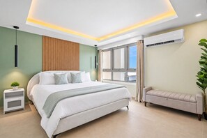 A modern bedroom with a large bed, bedside tables, a bench, and an air conditioning unit. - Remodeled Isla Verde Condo • One block to Beach (Carolina)