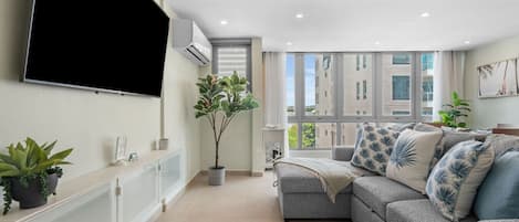 A modern living room with a grey sofa, a flat-screen TV mounted on the wall, a potted plant, and a large window with a view of buildings.