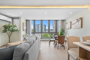 Ein modernes Wohnzimmer mit einer Couch, einem Esstisch, Stühlen und einem großen Fenster mit Blick auf Gebäude und Grünflächen.