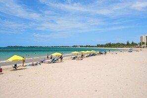 Ein Strand mit Liegestühlen und Sonnenschirmen, ein klarer Himmel und eine entfernte Küstenlinie.