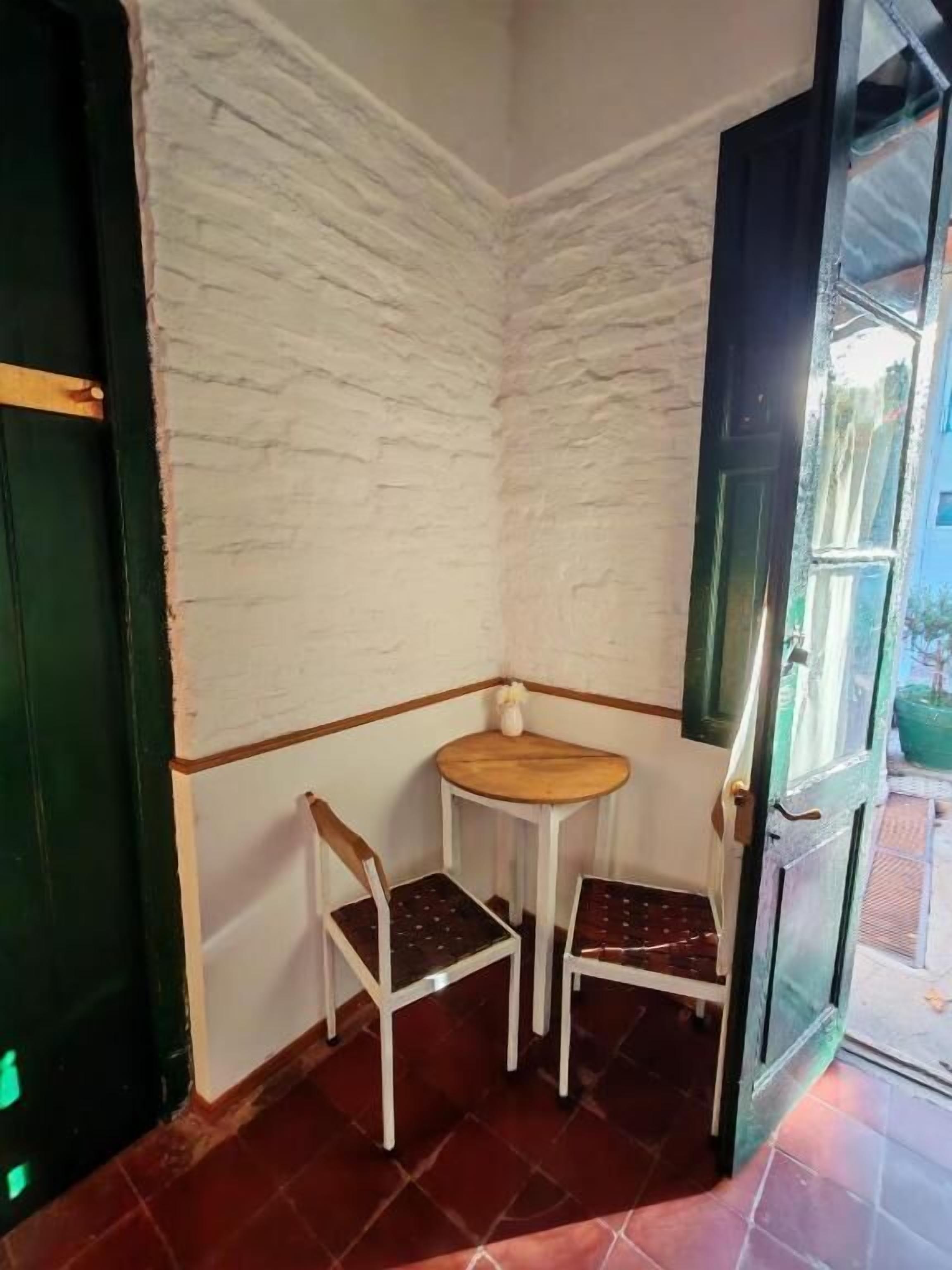 Una habitación de esquina con una pared blanca texturizada, una pequeña mesa de madera, dos sillas blancas y una puerta verde.