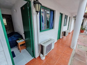 Een gang met een rij kamers, elk met een raam en een deur. Er staat een klein tafeltje met een plantje vlakbij de ingang van een kamer.