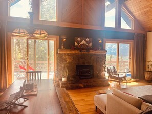 A spacious living area with a stone fireplace, wooden flooring, and large windows offering a view of the outdoors.