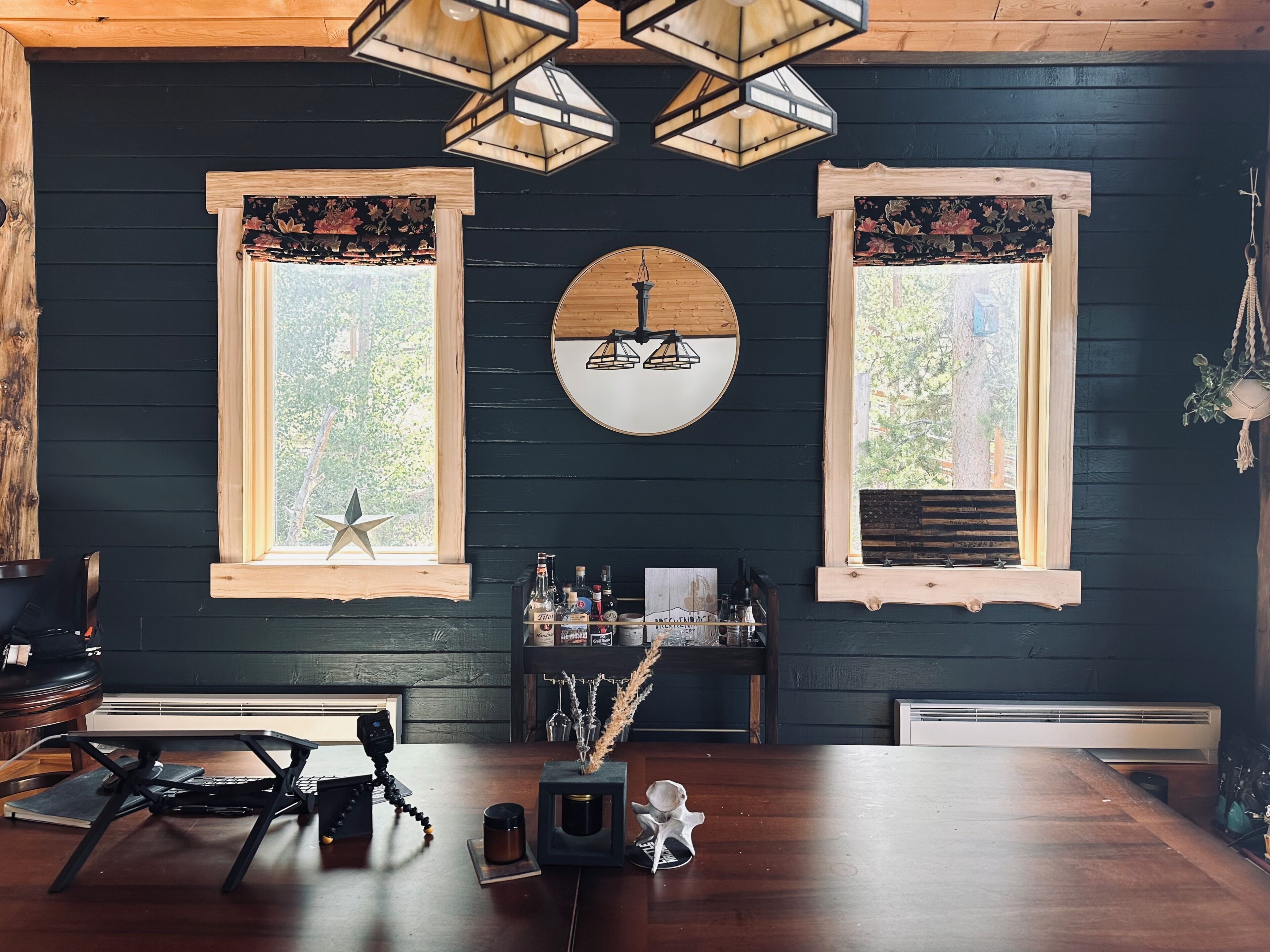 Une pièce avec un mur en bois foncé, deux fenêtres ornées de rideaux à motifs floraux, un miroir rond et une table en bois sur laquelle est posé un appareil photo sur trépied.