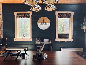 A room with a dark wooden wall, two windows with floral curtains, a round mirror, and a wooden table with a tripod camera.
