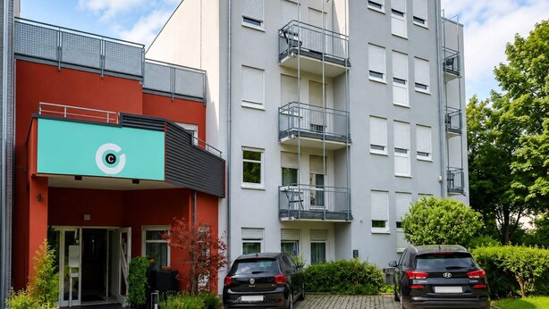 A multi-story residential building with a red facade, a blue sign with a white 'C' logo, and two parked cars in front. - 4 Person Holiday Park Home in Grossbettlingen (Grossbettlingen)