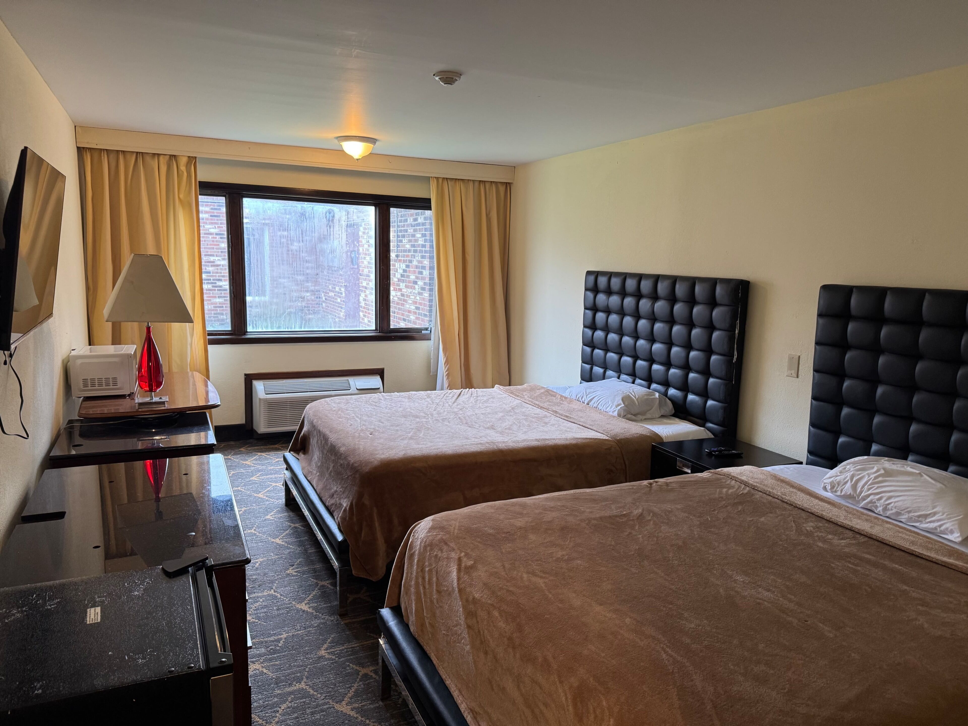 Habitación de hotel con dos camas, un escritorio, una lámpara y un ventanal con cortinas.