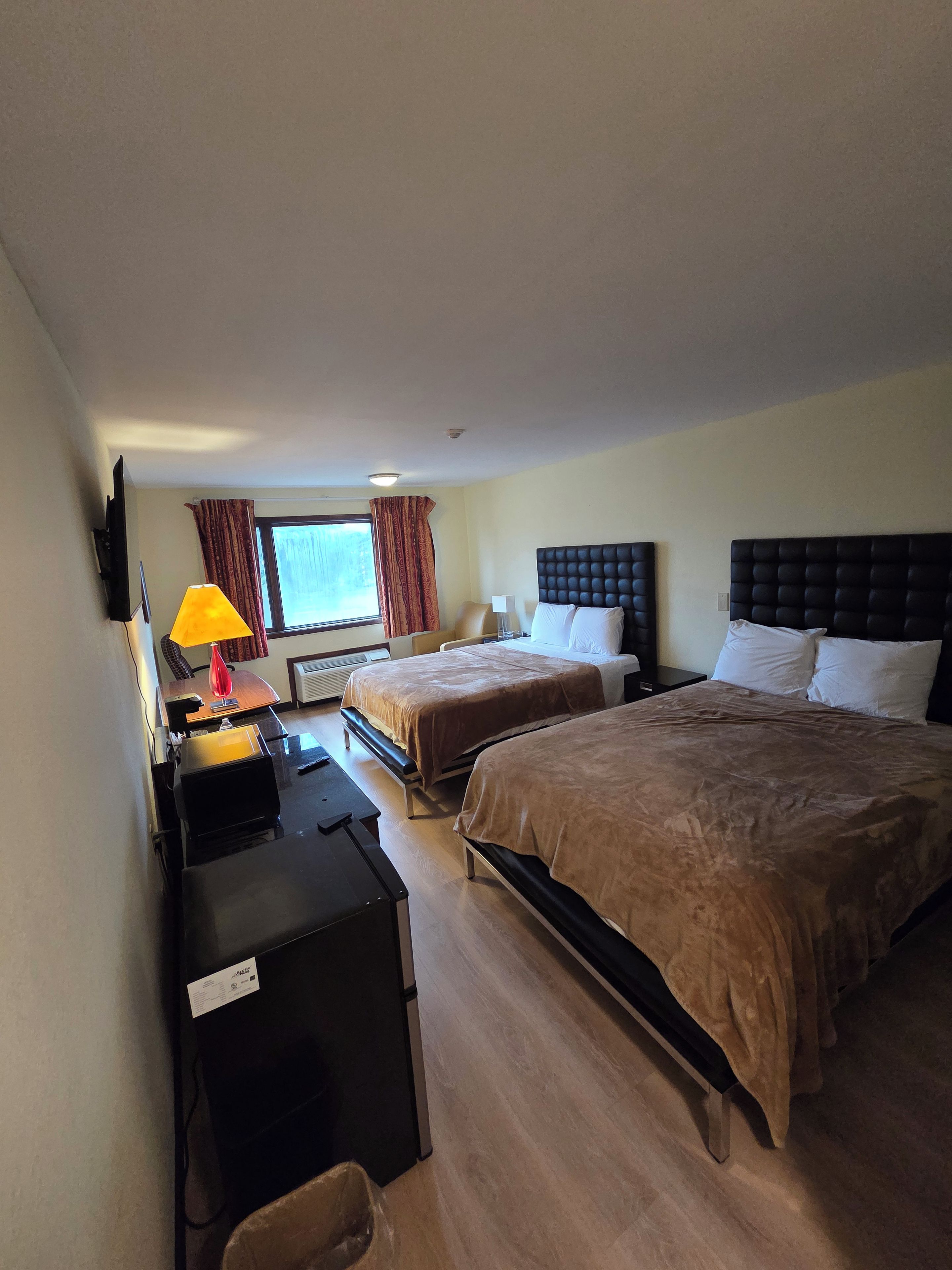 Una camera d'albergo con due letti, una scrivania, una sedia, una lampada e una finestra con tende.