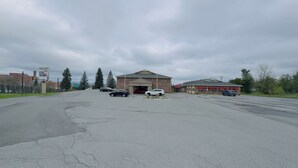 A parking lot with several cars, a building with a sign for Hilltop Inn, and a fenced area with trees. - HILLTOP INN (Barre)