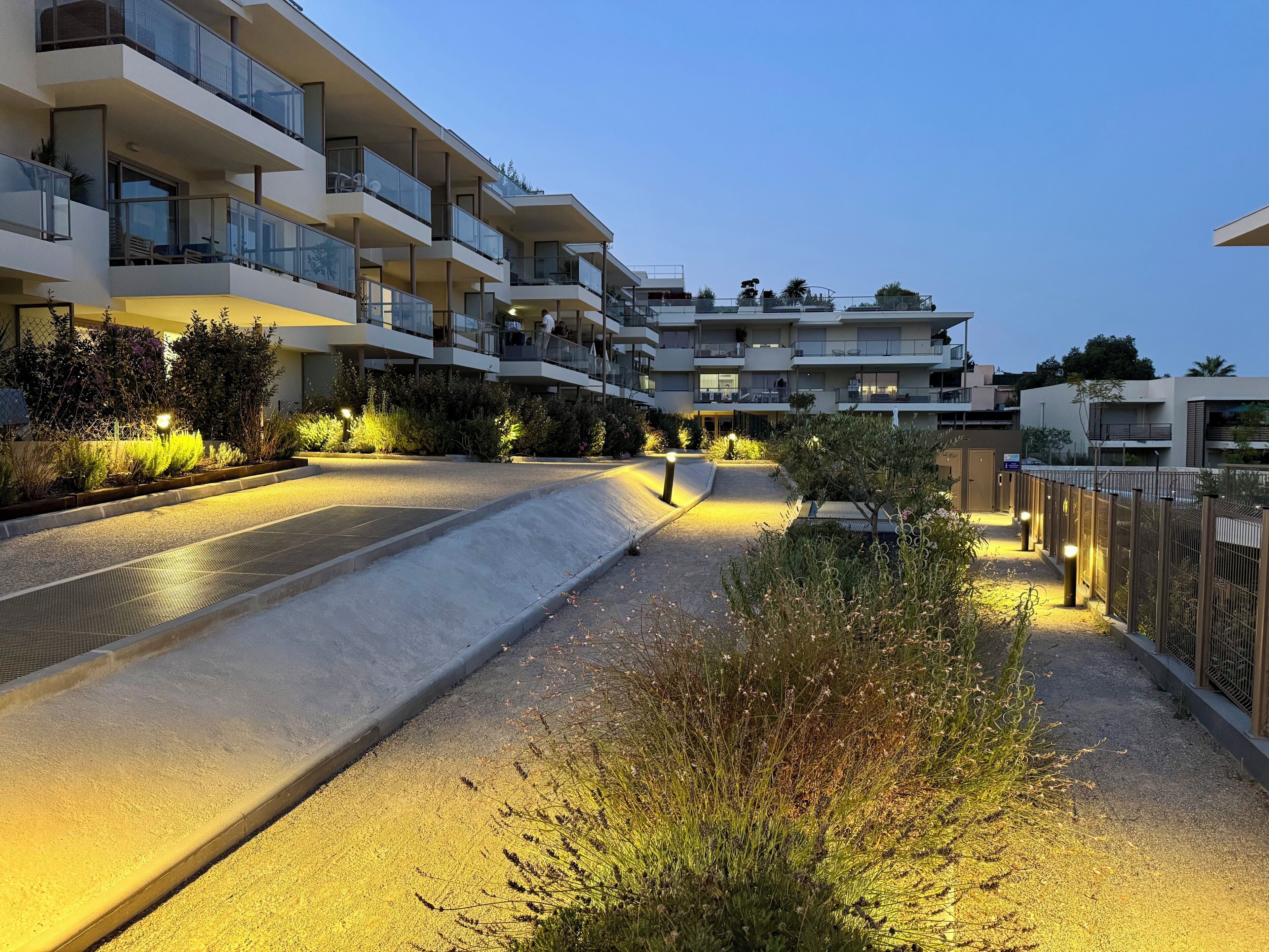A multi-story residential building with balconies, a landscaped area with plants, and a paved walkway.