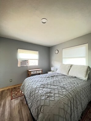 A bedroom with a bed, a nightstand, a wall clock, a window with blinds, and a ceiling light.