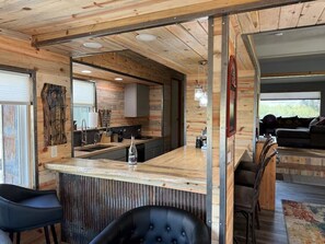 A rustic interior with a wooden bar area, seating, and a kitchen visible through an open doorway.