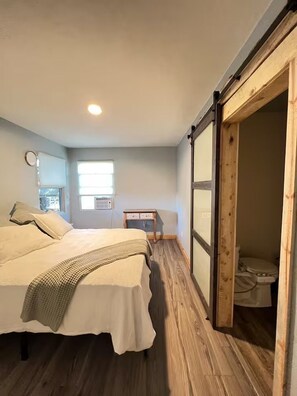 Une chambre à coucher comprenant un lit, une table de chevet, une fenêtre et une porte en bois donnant sur une salle de bain.