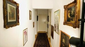 A hallway with framed artwork on the walls, a wooden floor, and a black lamp on the right.