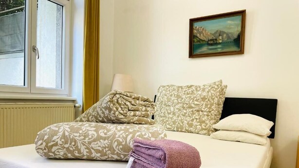 A neatly made bed with patterned pillows and a folded towel, a painting on the wall, and a window with curtains.