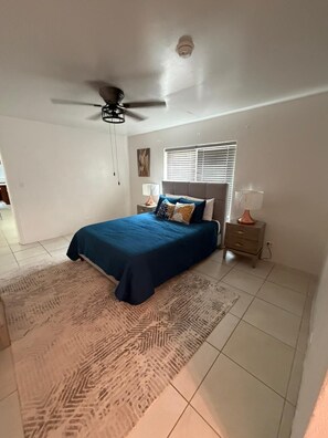 A bedroom with a bed, a ceiling fan, a window with blinds, a nightstand, and a rug.