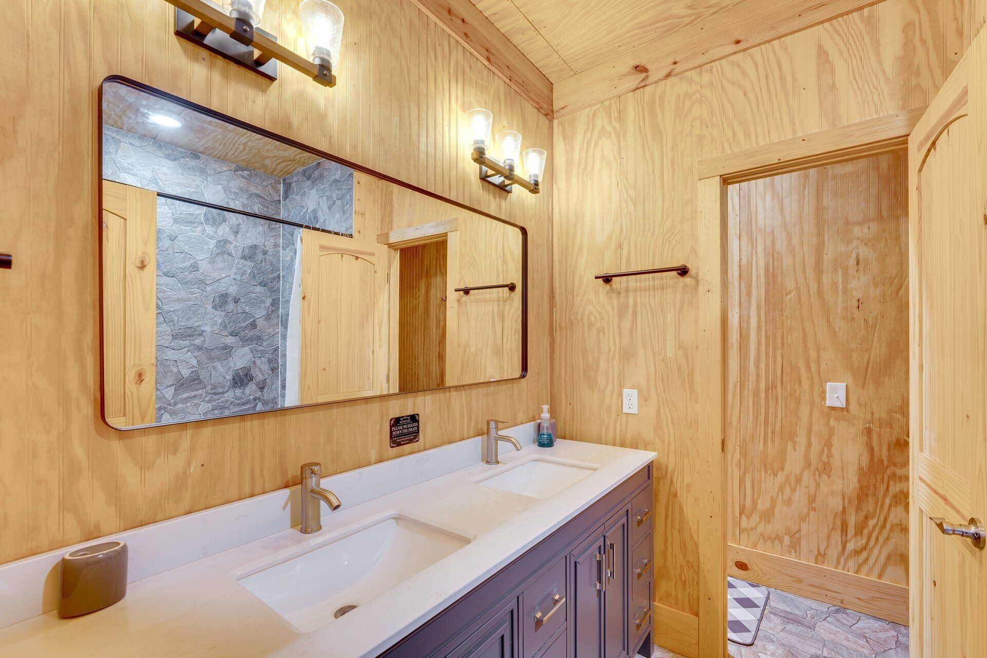 Un baño con paredes de madera, un espejo grande, lavamanos doble y una puerta con toallero.