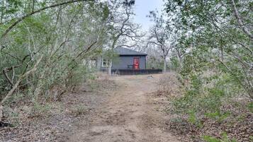 Une cabane avec une porte rouge, entourée d’une forêt dense et accessible par un sentier de terre.