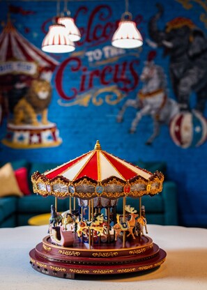 A carousel with horses and riders, set against a mural with circus-themed artwork. - Hollyhock Suite 313: 5 beds/2 bath (Louisville)