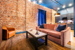 A modern living room with a brick wall, a sofa, a coffee table, and a kitchen area in the background. - Hollyhock Suite 310: 2 beds/1 bath (Louisville)