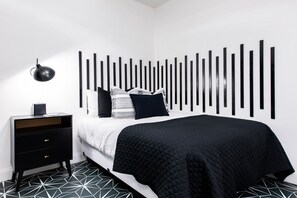 Ein modernes Schlafzimmer mit einem schwarz-weiß gestreiften Kopfteil, einem schwarzen Nachttisch mit einer Lampe und einem Boden mit geometrischen Mustern.