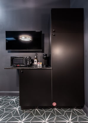 A black refrigerator with a built-in microwave and coffee maker, a flat-screen TV mounted above it, and a geometric patterned floor. - Hollyhock Suite 309: 1 bed/1 bath (Louisville)