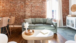 Ein Wohnzimmer mit Backsteinwand, grauem Sofa, weißem Tisch, Korbstühlen und einem weißen Schrank.