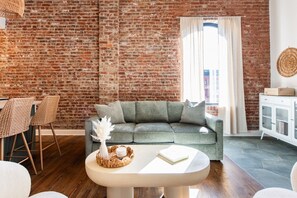 A living room with a brick wall, a grey sofa, a white table, wicker chairs, and a white cabinet. - Hollyhock Suite 307: 3 bed /2 bath (Louisville)