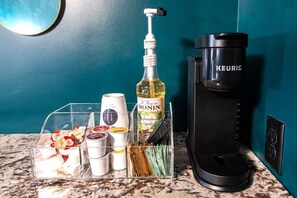 A kitchen counter with a coffee maker, a bottle of Monin French Vanilla syrup, and a clear plastic organizer containing various condiments and utensils. - Hollyhock Suite 311: 1 bed/1 bath (Louisville)