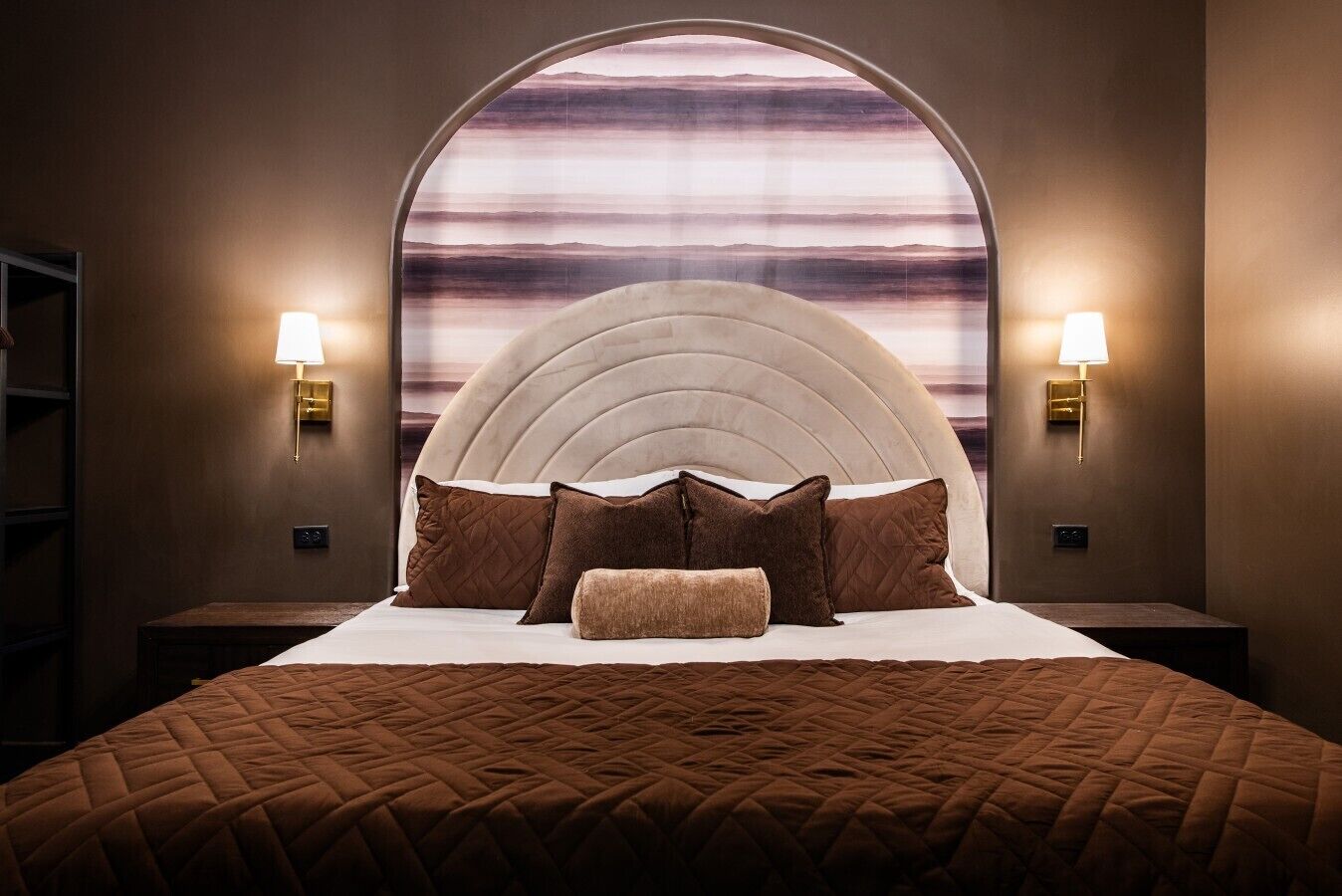 A bedroom with a large bed, a headboard with a curved design, two bedside lamps, and a view through a window.
