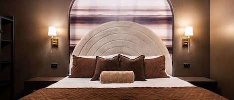 Ein Schlafzimmer mit einem großen Bett, einem Kopfteil mit geschwungener Form, zwei Nachttischlampen und Blick durch ein Fenster.