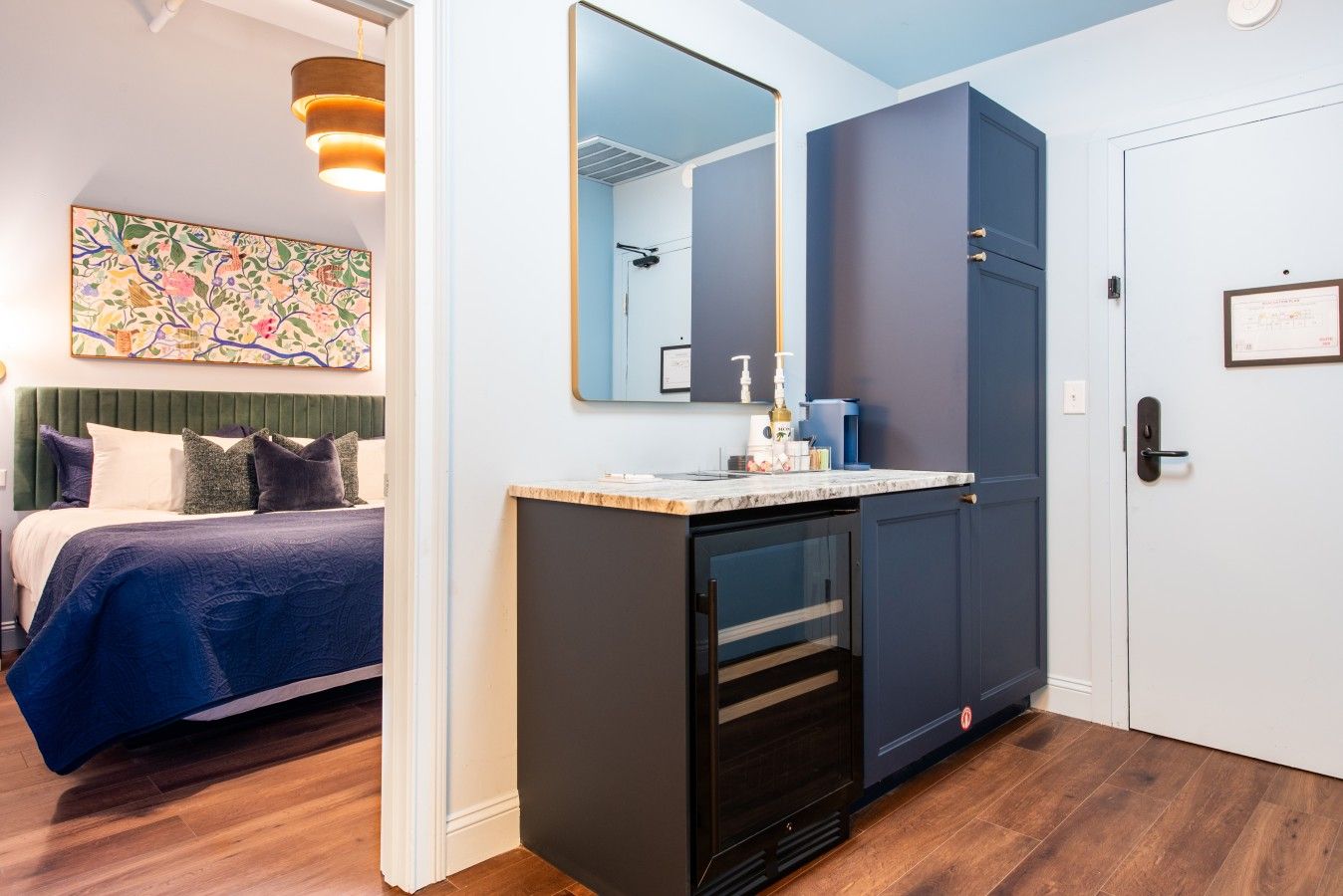 A hotel room with a bed, a desk with a mirror, a door, and a wall with a framed certificate.