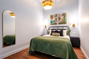 A bedroom with a bed, a nightstand, a wall-mounted mirror, and two wall-mounted lights. - Hollyhock Suite 308: 2 beds/1 bath (Louisville)