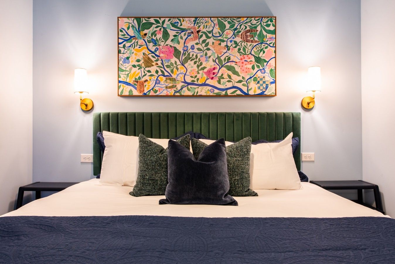 A hotel room with a bed, pillows, a headboard, a wall-mounted artwork, and two wall sconces.