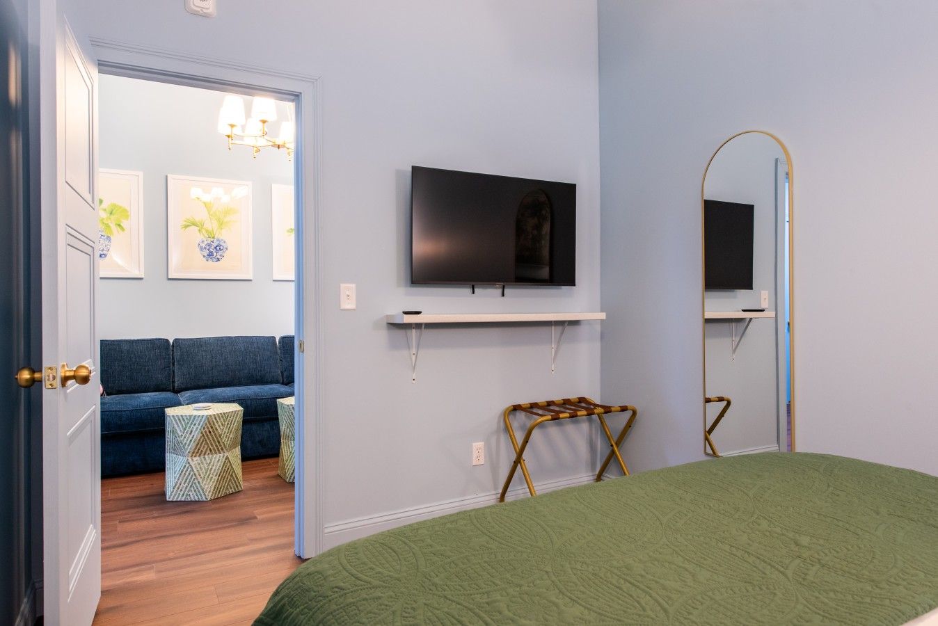 A bedroom with a bed, a television mounted on the wall, a mirror, and a door leading to a living area with a sofa and a chandelier.