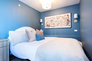 A bedroom with a bed, pillows, a nightstand, a wall-mounted lamp, and a floral artwork. - Hollyhock Suite 306: 1 bed + 1 loft bed/1 bath (Louisville)