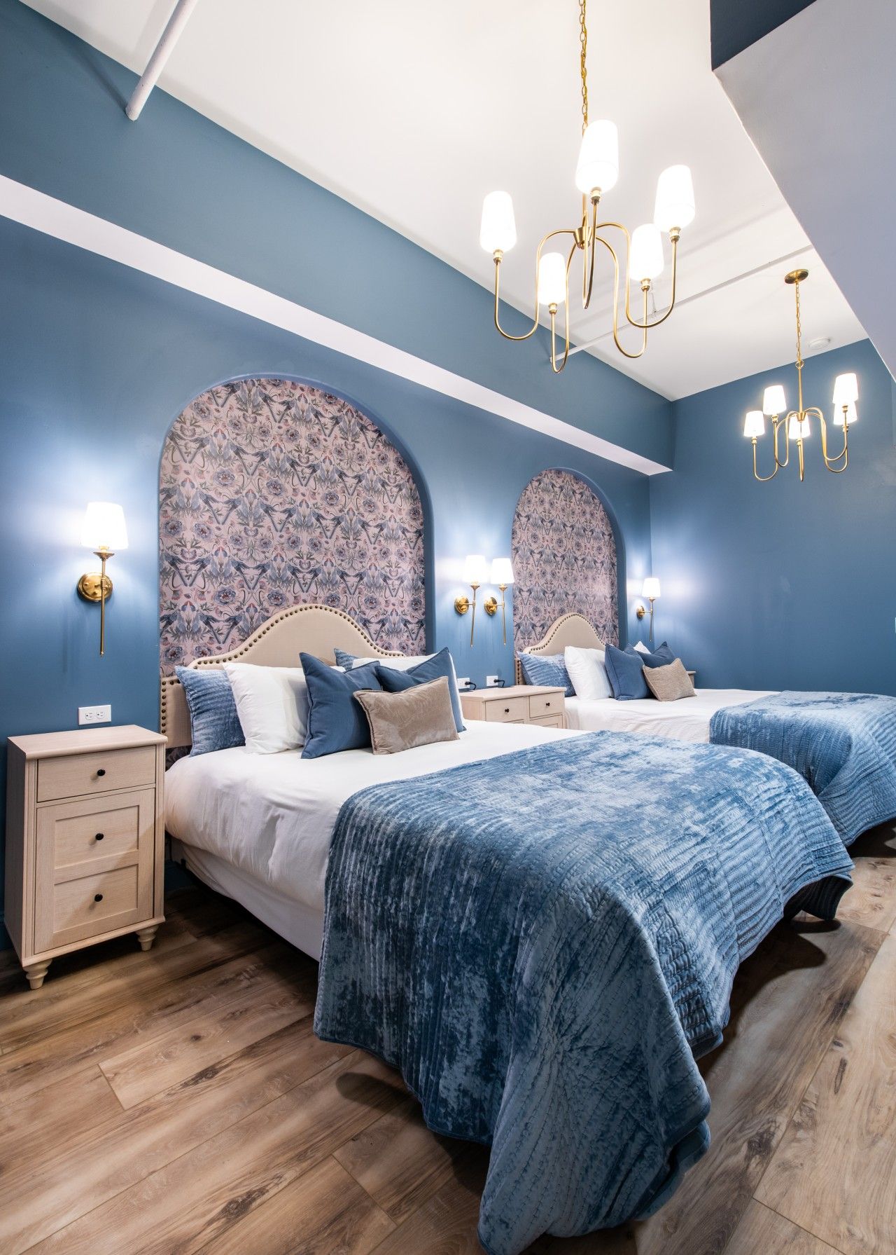 A bedroom with two beds, a chandelier, patterned wallpaper, and a wooden floor.