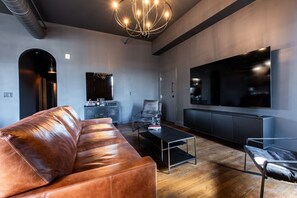 A modern living room with a leather sofa, a coffee table, a TV, and a chandelier. - Hollyhock Suite 502: 4 bed/2 bath with soaking tub (Louisville)