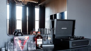 Un coin bar avec un miroir, un lustre et un haut-parleur. On y trouve des verres, des bouteilles et un livre sur le comptoir.