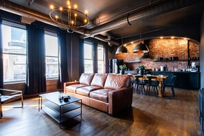 A modern living room with a leather sofa, a dining table, and a kitchen area with exposed brick walls and industrial lighting. - Hollyhock Suite 502: 4 bed/2 bath with soaking tub (Louisville)
