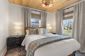 A bedroom with a wooden ceiling, a bed with patterned bedding, two bedside tables with lamps, and a view of the outdoors through large windows. - Fall Getaway with Gas Fire Table | Walk to Lake, Restaurants | Sleeps 12 (Williams Bay)