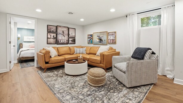 Ein modernes Wohnzimmer mit einem braunen Ledersofa, einem gestreiften Sessel und einem runden Hocker. An der Wand befinden sich gerahmte Bilder, ein Fenster mit weißen Vorhängen und eine Tür zu einem anderen Raum.