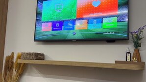 Ein flachbildfernseher an der Wand, darunter ein Holzregal mit Dekorationsobjekten und einer Vase mit Trockenblumen.