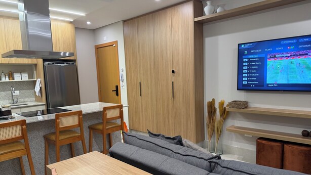 Ein modernes Wohnzimmer mit einer Couch, einem Esstisch und Stühlen. Es gibt eine Küchenzeile mit Kühlschrank und Schränken. Ein Fernseher an der Wand zeigt einen Sportkanal.