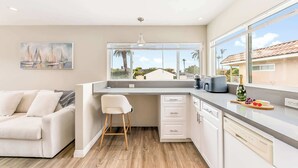 A modern kitchen with a large window, a dining area with a white sofa, and a painting on the wall. - Del Mar Studio - Walk to Beach (Del Mar)