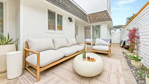 Un área de estar moderna al aire libre con sofá y sillas, una mesa redonda con una botella y fruta, y una planta en maceta.