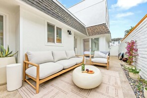 Ein moderner Außenbereich zum Sitzen mit einer Couch und Sesseln, einem runden Tisch mit einer Flasche und Früchten sowie einer Topfpflanze.