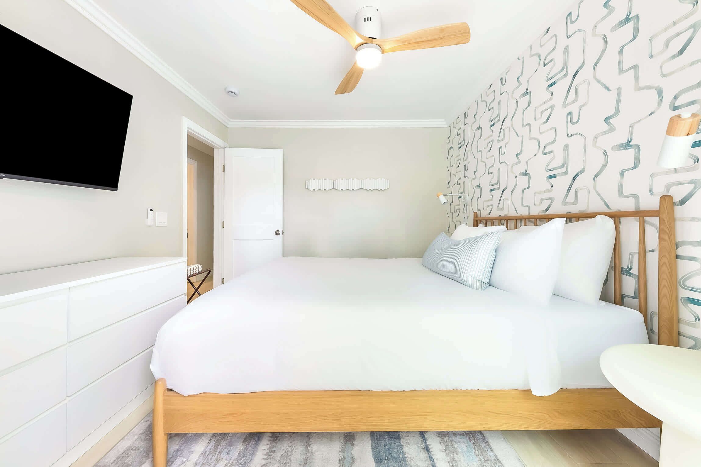 A bedroom with a bed, a ceiling fan, a TV mounted on the wall, and a patterned wall.