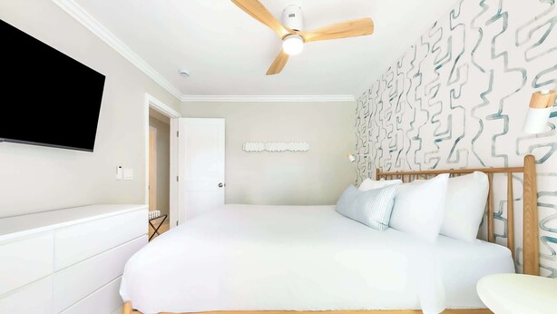 Un dormitorio con cama, ventilador de techo, televisor montado en la pared y un papel tapiz estampado.