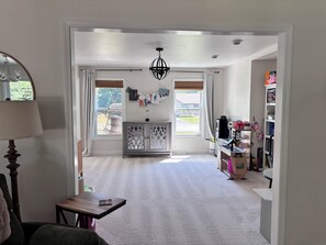 A spacious room with a high ceiling, a chandelier, a large window with curtains, a wooden cabinet, and a bookshelf. - Spacious 4-bedroom home less than 5 miles away from EAA in Oshkosh with a gym (Oshkosh)